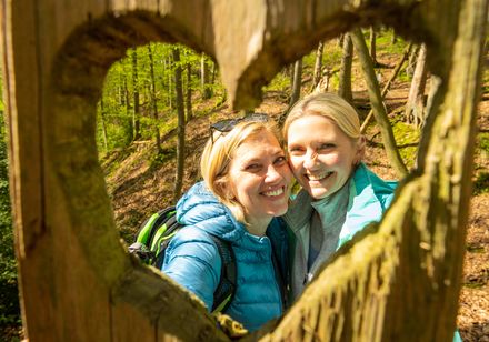 Zwei Frauen lächeln durch herzförmiges Holzfenster im Wald, umgeben von sonnigem Licht.