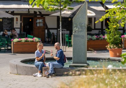 Zwei Personen sitzen plaudernd an einem Brunnen mit kleiner Skulptur in einer sonnigen Straße.