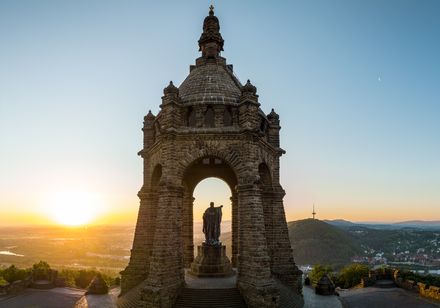 Denkmal mit Bismarck-Statue vor Sonnenuntergang, umgeben von sanften Hügeln und Flusstal.
