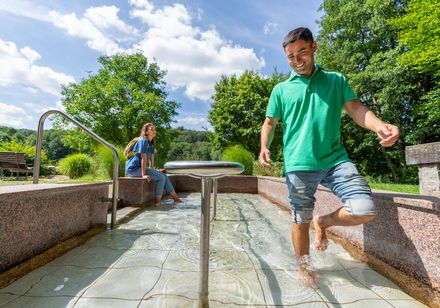 <p>Mann und Frau genießen ein Kneippbecken im Freien bei sonnigem Wetter und grüner Umgebung.</p>