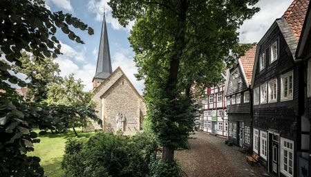 Historische Fachwerkhäuser umrahmen eine alte Kirche mit steilem Kirchturm auf einem grünen Platz.