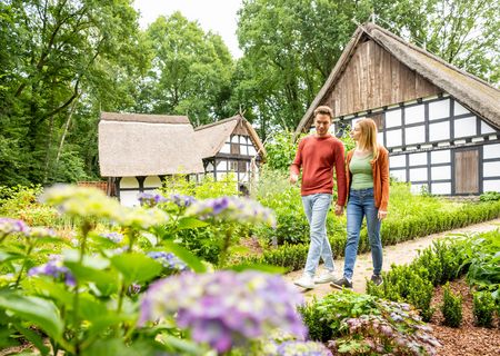 Ein junges Paar spaziert durch einen blühenden Garten vor Fachwerkhäusern in ländlicher Umgebung.