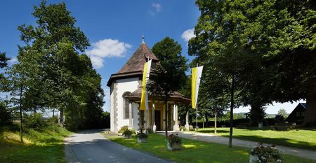 Annenkapelle mit spitzem Holzdach und gelben Fahnen, umgeben von Bäumen, neben einer Straße.