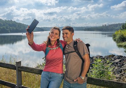Paar macht Selfie vor idyllischem See mit bewaldeten Ufern und bewölktem Himmel im Hintergrund.