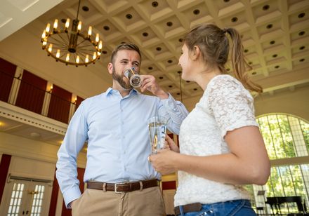 Zwei Personen stehen in einem elegant gestalteten Raum mit Kronleuchter, lächeln und trinken Wasser.