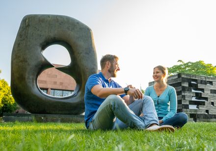 <p>Ein Paar sitzt lächelnd auf der Wiese vor einer großen, modernen Steinskulptur im Park.</p>