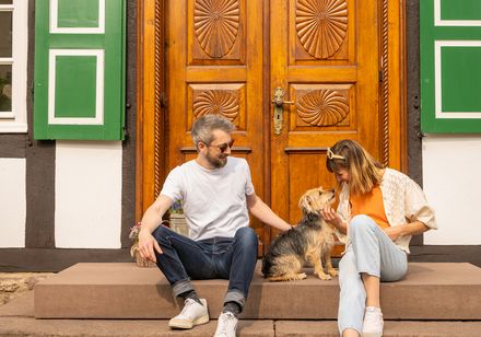 Ein Paar mit Hund sitzt entspannt auf Stufen vor einem Fachwerkhaus mit grünen Fensterläden.