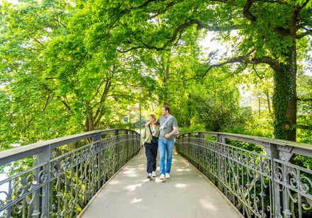 Zwei Menschen spazieren bei sonnigem Wetter über eine Brücke im grünen Aawiesenpark Herford.