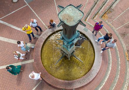 Menschen stehen um einen runden Brunnen mit bronzener Skulptur, auf gemustertem Pflaster.