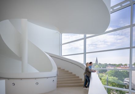 Zwei Menschen stehen auf einer modernen Wendeltreppe mit Glasfront und blicken auf eine Kirche.
