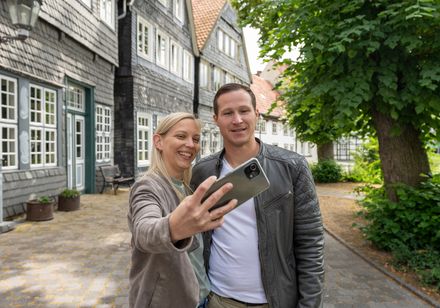 Paar macht Selfie vor Fachwerkhäusern in malerischer gepflasterter Straße bei grünen Bäumen.