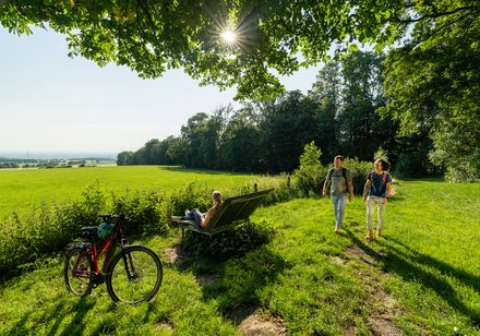 Drei Menschen im Grünen bei Sonnenschein, eine sitzt auf Bank, zwei gehen, Fahrrad im Vordergrund.