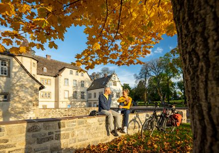 Ein Paar sitzt unter goldenem Herbstlaub vor einem historischen Gebäude, redet und genießt die Sonne.