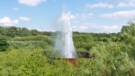 Wasserskulptur Aqua Magica in einem bewaldeten Park, umgeben von üppigem Grün unter blauem Himmel.