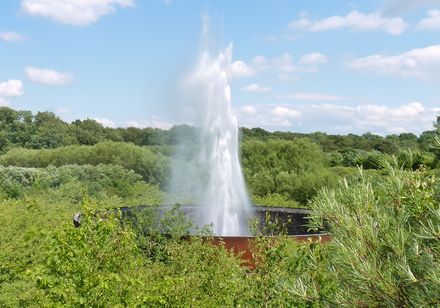 Wasserskulptur Aqua Magica in einem bewaldeten Park, umgeben von üppigem Grün unter blauem Himmel.