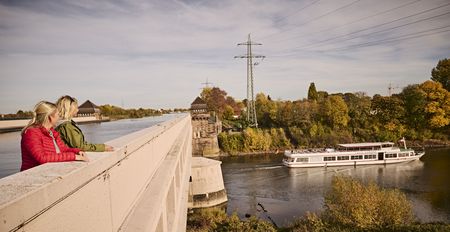 Zwei Frauen stehen auf einer Brücke und blicken auf ein vorbeifahrendes Ausflugsschiff im Herbst.