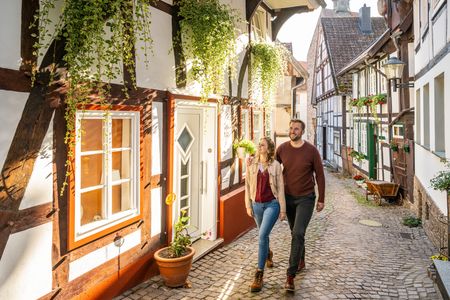 Ein Paar spaziert durch eine malerische historische Gasse mit Fachwerkhäusern und blühenden Pflanzen.