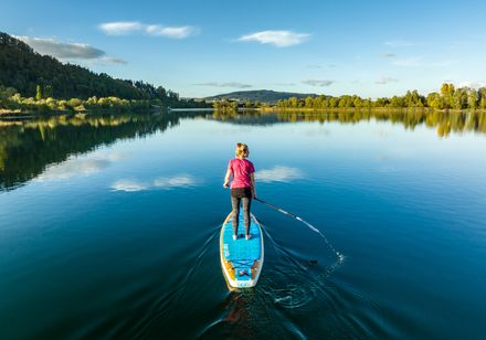 Person steht auf einem blauen Stand-up-Paddle-Board mitten auf einem ruhigen, spiegelnden See.