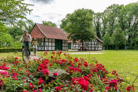 Zwei Radfahrer fahren an einem Fachwerkhaus in einer grünen Parklandschaft mit roten Blumen vorbei.