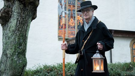 Ein Nachtwächter in traditionellem Gewand mit Laterne und Hellebarde vor einer historischen Kirche.