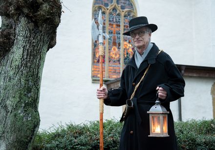 Ein Nachtwächter in traditionellem Gewand mit Laterne und Hellebarde vor einer historischen Kirche.