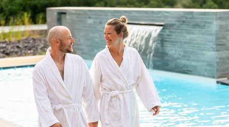 Das Bild zeigt ein Paar im Bademantel vor einem modernen Außenpool mit Wasserfall.