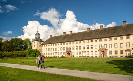 Zwei Personen spazieren vor der barocken Fassade von Schloss Corvey bei sonnigem Wetter.
