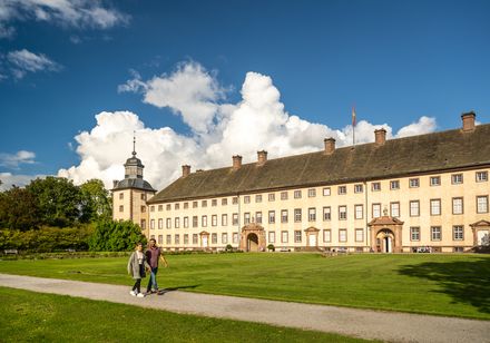 Zwei Personen spazieren vor der barocken Fassade von Schloss Corvey bei sonnigem Wetter.