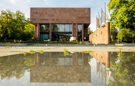 <p>Moderne Architektur mit Glasfront spiegelt sich in einer Pfütze, umgeben von Bäumen und blauem Himmel.</p>