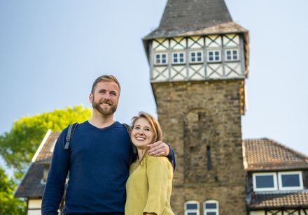 Junges Paar steht lächelnd vor einem historischen Gebäude mit Fachwerk und Turm unter blauem Himmel.