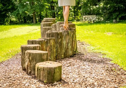 Person geht barfuß über Holzstämme im Barfußpfad im Kurpark Bad Holzhausen, umgeben von Natur.