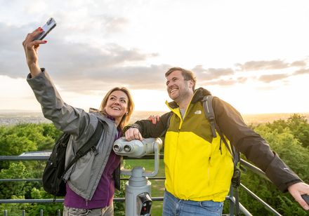 Zwei Menschen machen ein Selfie auf einem Aussichtsturm, umgeben von grüner Landschaft im Sonnenuntergang.