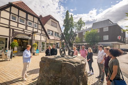 Eine Gruppe beobachtet eine Skulptur mit Schlüsseln auf einem großen Stein im sonnigen Dorfzentrum.