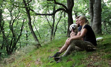 Zwei Personen sitzen im Wald auf einer Moosfläche, begleitet von einem kleinen, weißen Hund.