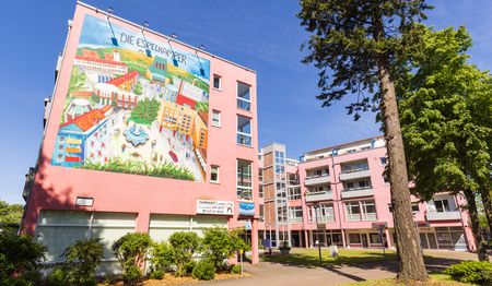 Großes Wandgemälde an einem rosa Wohngebäude, Bäume im Vordergrund, blauer Himmel.