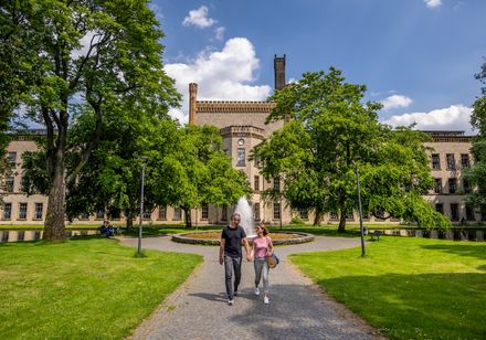Ein Paar spaziert auf einem gepflasterten Weg durch eine grüne Allee vor einem historischen Gebäude.