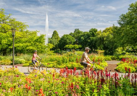 Ein Paar radelt durch einen farbenfrohen Park mit blühenden Blumen und einem hohen Wasserbrunnen.