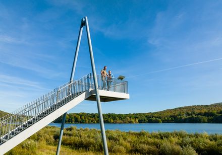 Metallische Aussichtsplattform mit zwei Personen, bietet Blick über einen See und bewaldete Hügel.