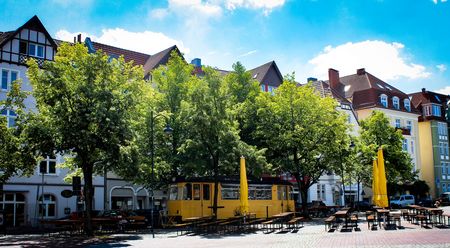 Gelbe Straßenbahn in belebtem Stadtplatz mit Bäumen und Cafés, umgeben von historischen Gebäuden.