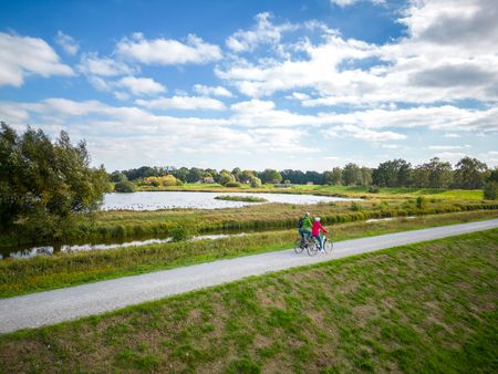 Radfahrer auf einem ländlichen Weg neben einem Teich unter blauem Himmel mit weißen Wolken.