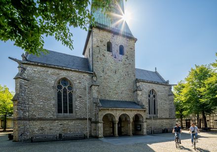 Fahrradfahrer vor historischer Kirche mit hohem Turm unter klarem, sonnigem Himmel, umgeben von Bäumen.