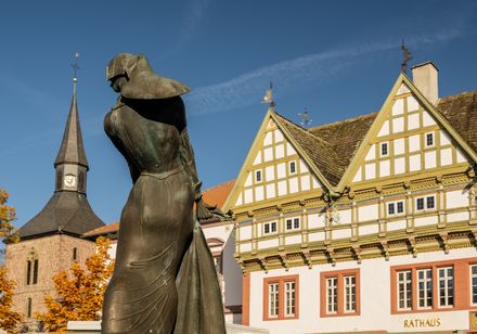 <p>Bronzestatue in Altstadt vor Fachwerkhäusern und Kirchturm, Bäume im Herbstlaub, blauer Himmel.</p>