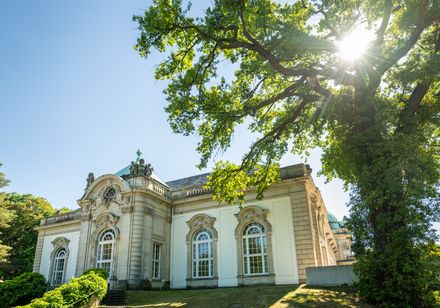 Historisches Gebäude mit verzierten Fenstern, umgeben von grünem Garten und strahlendem Sonnenlicht.