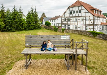 Zwei Menschen sitzen auf einer überdimensionalen Holzbänkenkulisse vor einem Fachwerkhaus mit Garten.