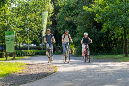 Drei Menschen fahren bei sonnigem Wetter auf einem kurvigen Pfad durch einen grünen Park Fahrrad.