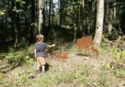 Ein kleiner Junge betrachtet mit Interesse eine Wildschwein-Familie aus Metallskulpturen im Wald.