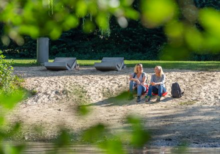 Zwei Frauen sitzen in Freizeitkleidung am sandigen Ufer eines ruhigen Sees, umgeben von Natur.