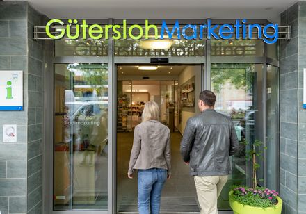 Ein Paar betritt das moderne Besucherzentrum von Gütersloh Marketing mit Infostand und Prospekten.