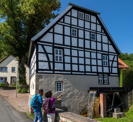 Fachwerkhaus mit Wasserrad, zwei Personen davor. Historische Architektur neben Straße und Bäumen.