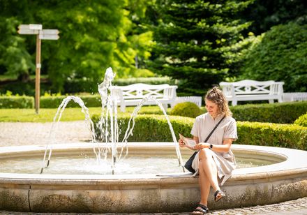 Frau liest Broschüre sitzend am Brunnen im Kurpark Bad Holzhausen